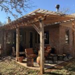Outdoor Living Space With A Wooden Pergola Comfortable Seating And A Stone Surface Clear Skies And Seasonal Leaves On The Ground