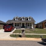 house with gray shingle roof green lawn and red sedan parked in driveway clear blue sky visible in background